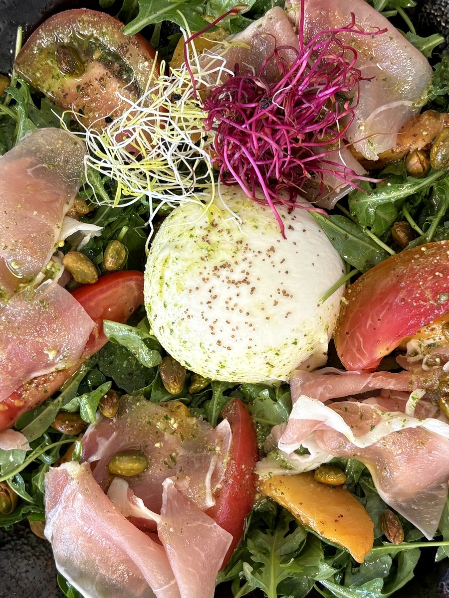 Burrata crémeuse entourée de jambon cru, tomates anciennes, pêches rôties, roquette et pistaches
